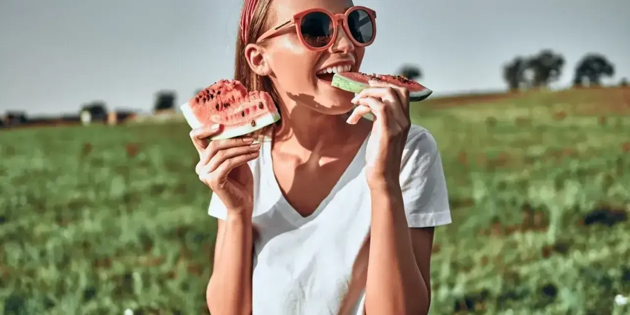 Dziewczyna w okularach zajada soczyste kawałki arbuza na tle zielonego pola. To idealny moment, kiedy jest sezon na arbuzy!
