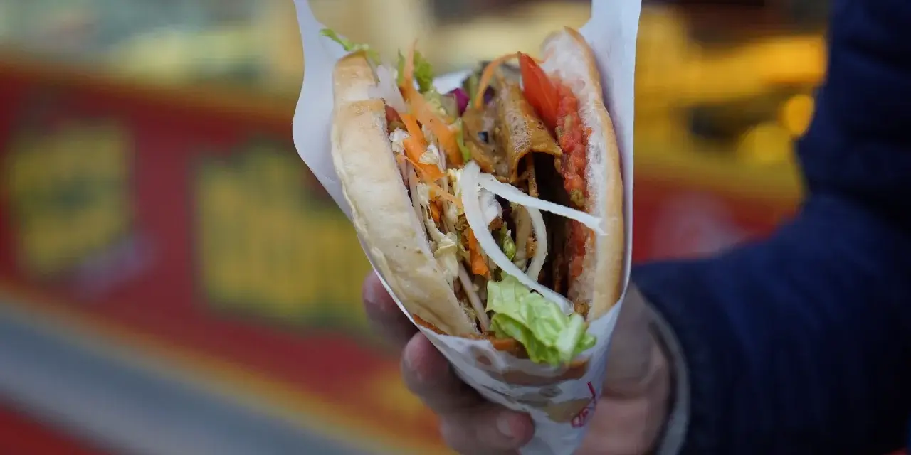Kebab w bułce z warzywami i mięsem, trzymany w dłoni.