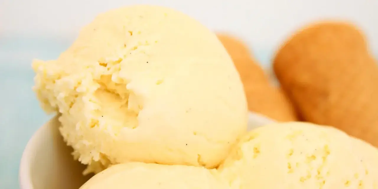 Cremoso helado de vainilla con motas oscuras, servido en un tazón blanco. Al fondo, conos de helado dorados esperan.