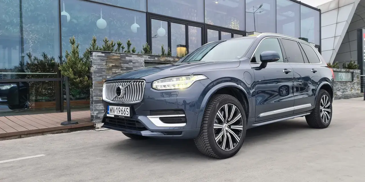 Elegancki, ciemnoszary Volvo XC90 T8 Recharge z polskimi tablicami rejestracyjnymi stoi przed nowoczesnym budynkiem z dużymi oknami.