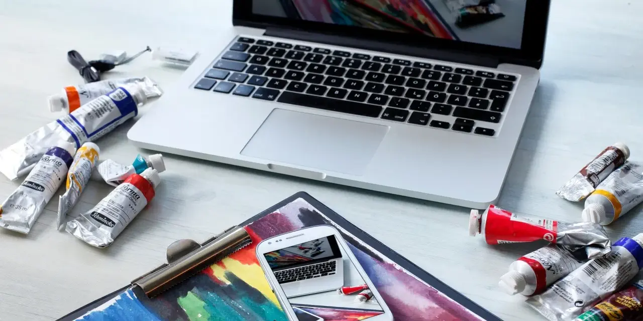 Na biurku leżą farby i laptop, na ekranie którego widać podobny obraz. Zastanawiasz się, ile zarabia grafik komputerowy?
