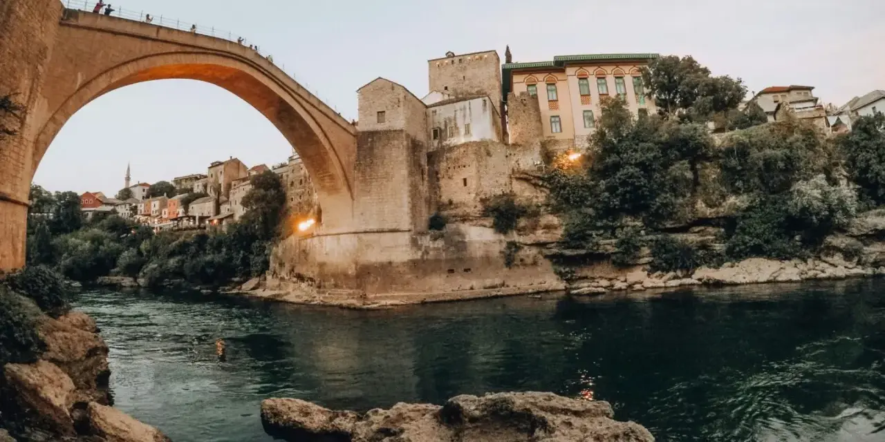 Iconic Stari Most bridge over the Neretva River, a must-see for any Bosnia travel adventure. People watch from the bridge as swimmers enjoy the emerald waters below.