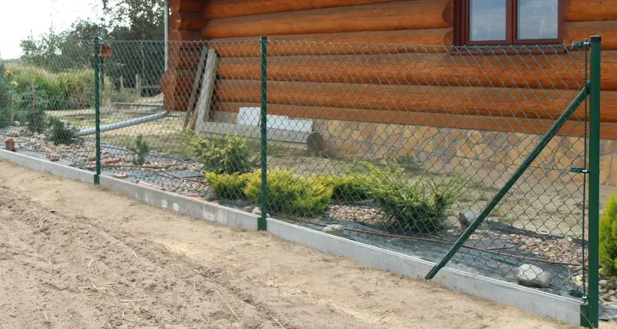 Montaż siatki ogrodzeniowej przy drewnianym domu. Widać zielone słupki, siatkę i betonowy podmurówkę.