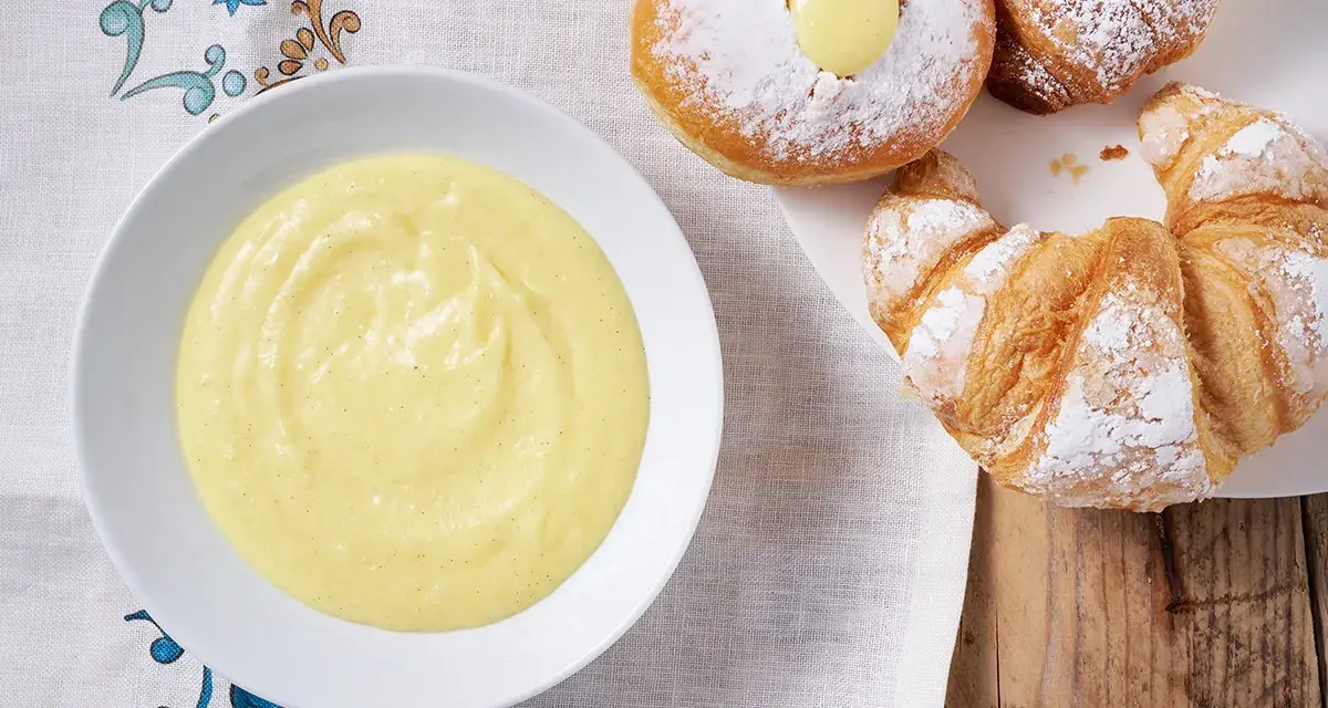 Una ciotola di soffice crema pasticcera senza uova accanto a cornetti e bomboloni spolverati di zucchero a velo.