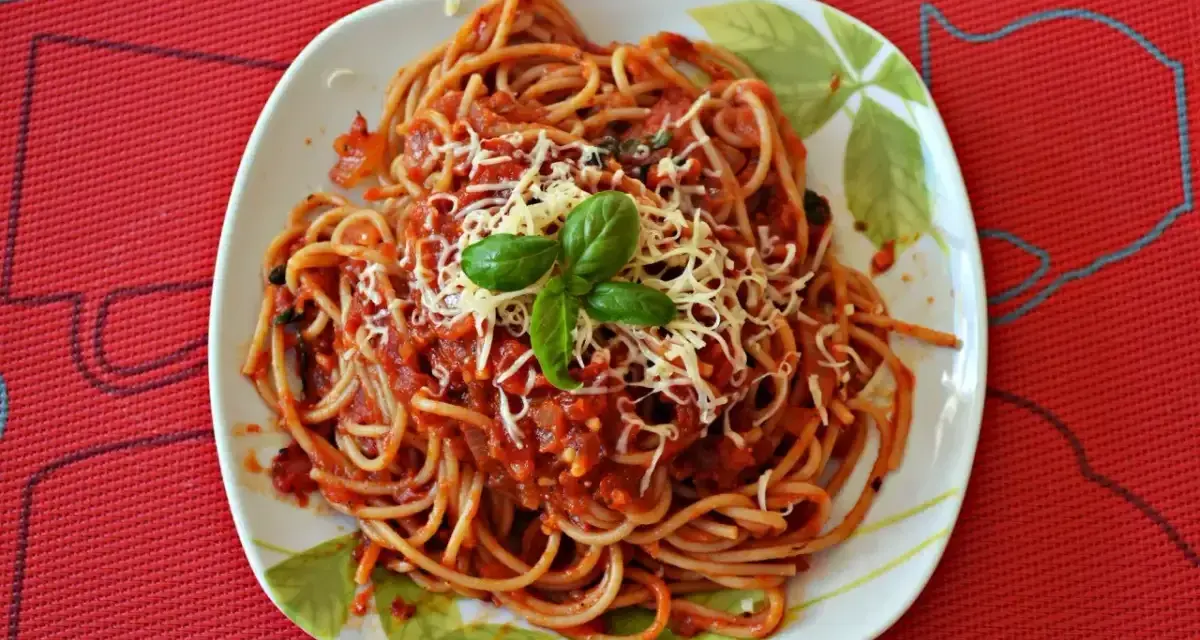 Pyszny sos do makaronu spaghetti, posypany startym serem i udekorowany listkiem bazylii, na talerzu z wzorem liści.