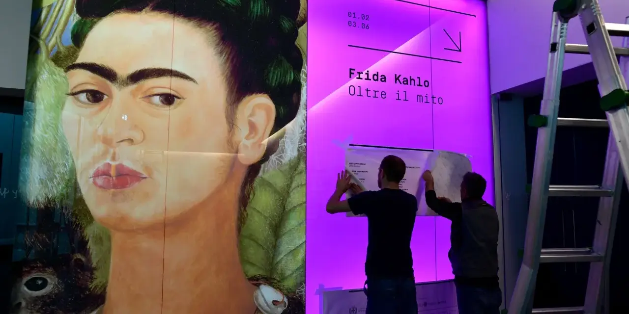 Un portrait de Frida Kahlo, avec deux hommes installant une affiche pour une exposition sur son histoire.