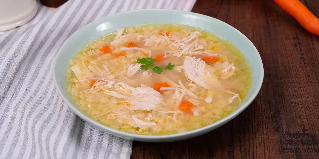 Un confortevole brodo di pollo con pezzi di pollo sfilacciato, carote e un rametto di prezzemolo, servito in una ciotola azzurra.