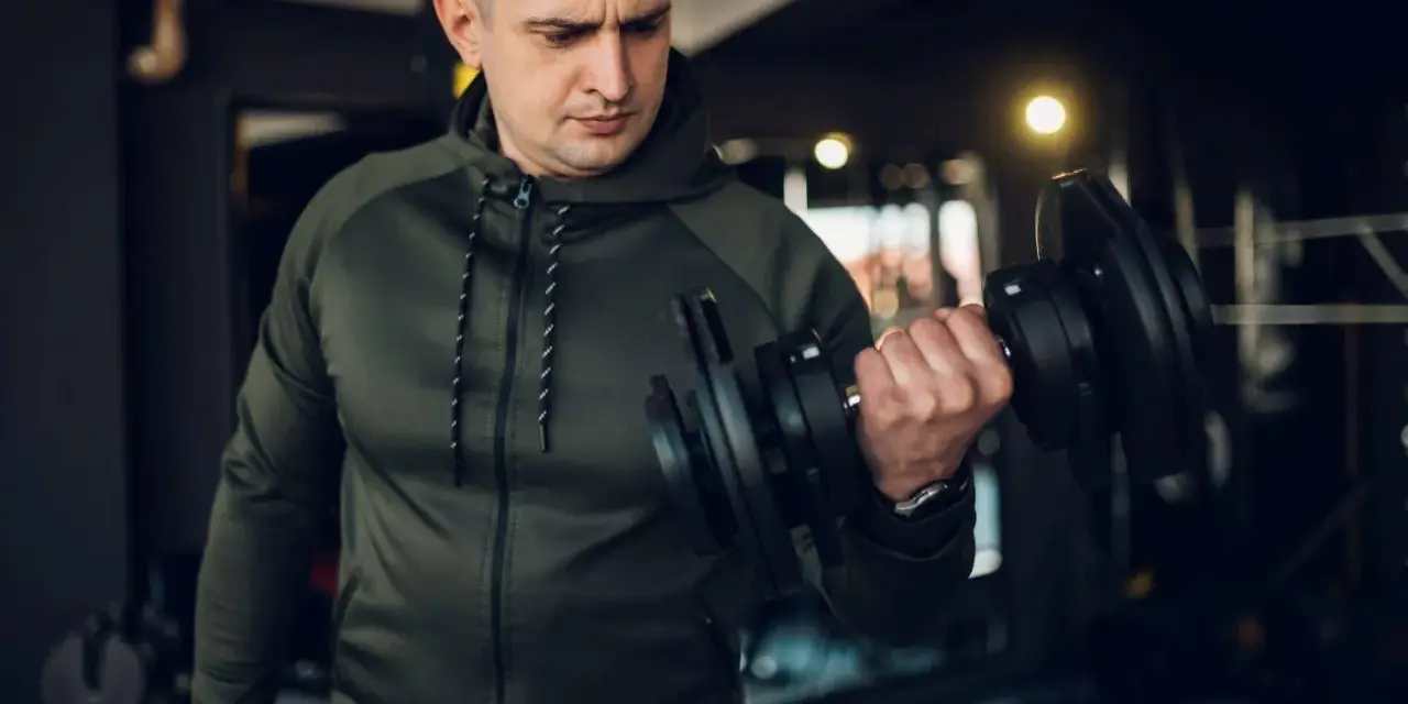 Mężczyzna w zielonej bluzie damskiej na siłownię ćwiczy biceps z hantlem.