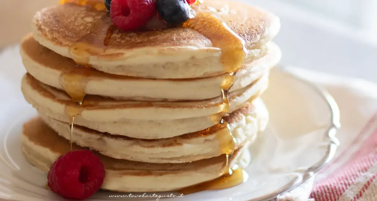 Pancakes senza uova dorati, impilati e inzuppati di sciroppo, guarniti con lamponi e mirtilli freschi.