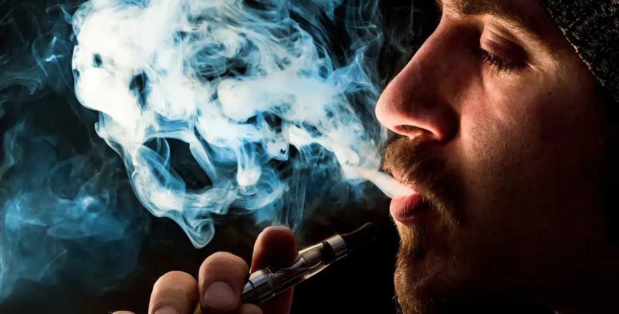 Hombre vapeando, el vaper sin nicotina es malo. El humo azul forma figuras fantasmagóricas.