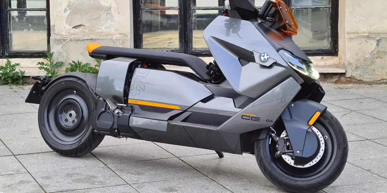 Szary skuter elektryczny BMW CE 04 z pomarańczowymi akcentami stoi na bruku przed budynkiem z dużymi oknami.