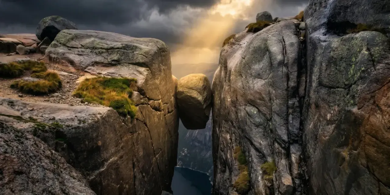 Enorm sten balanserar mellan klippor i Norge. Dramatiskt ljus bryter igenom molnen över en djup fjord.