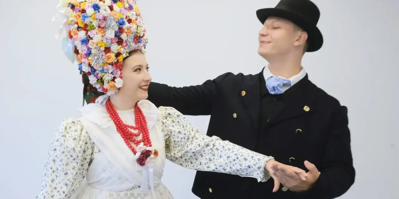 Para tańczy w strojach ludowych, co to folklor. Ona ma bogato zdobiony kwiatami wianek, on czarny kapelusz i marynarkę.