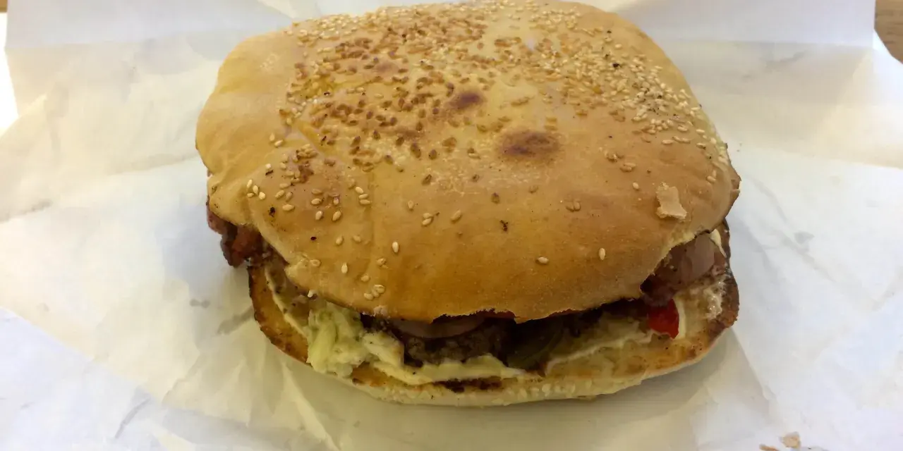 Burger z sezamem na białym papierze, widoczne składniki: mięso, sos, warzywa.