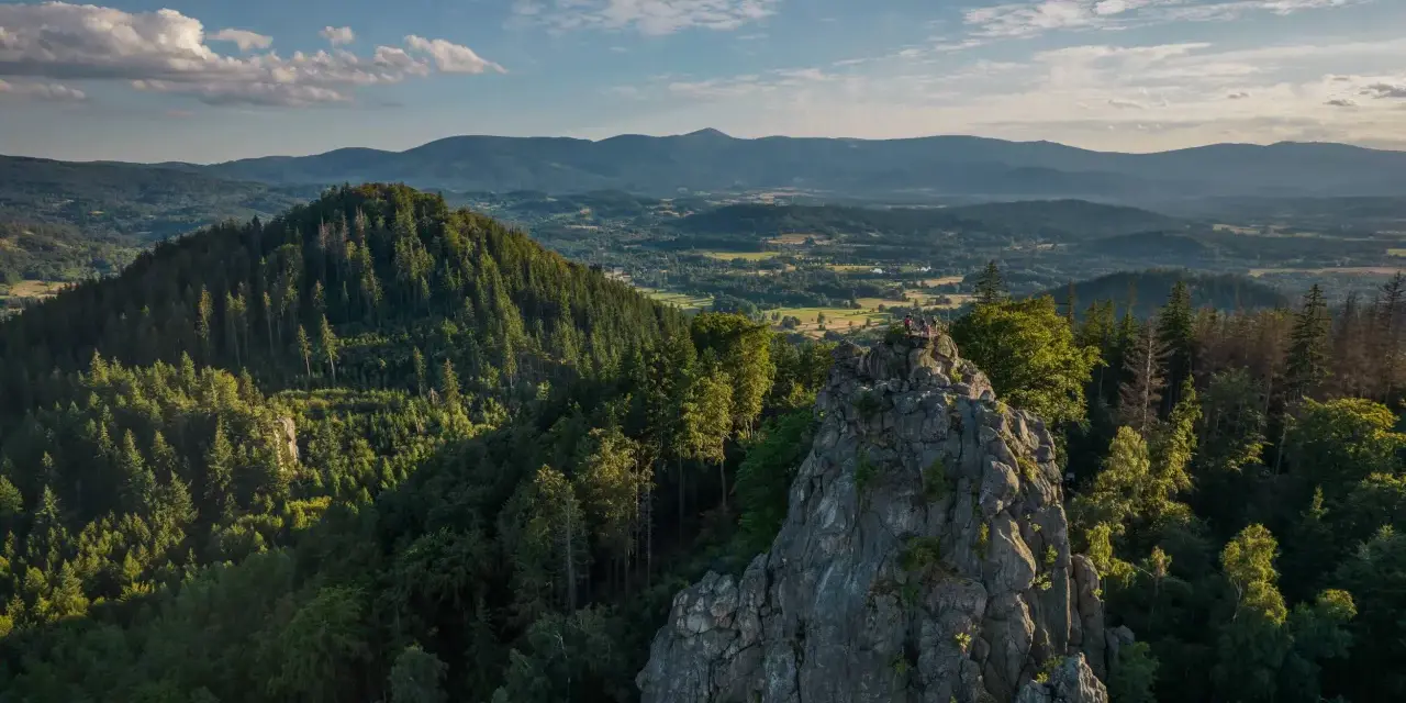 Widok na skalne formacje Rudawy Janowickie, porośnięte lasem, z ludźmi na szczycie. W tle rozległa panorama gór i dolin.