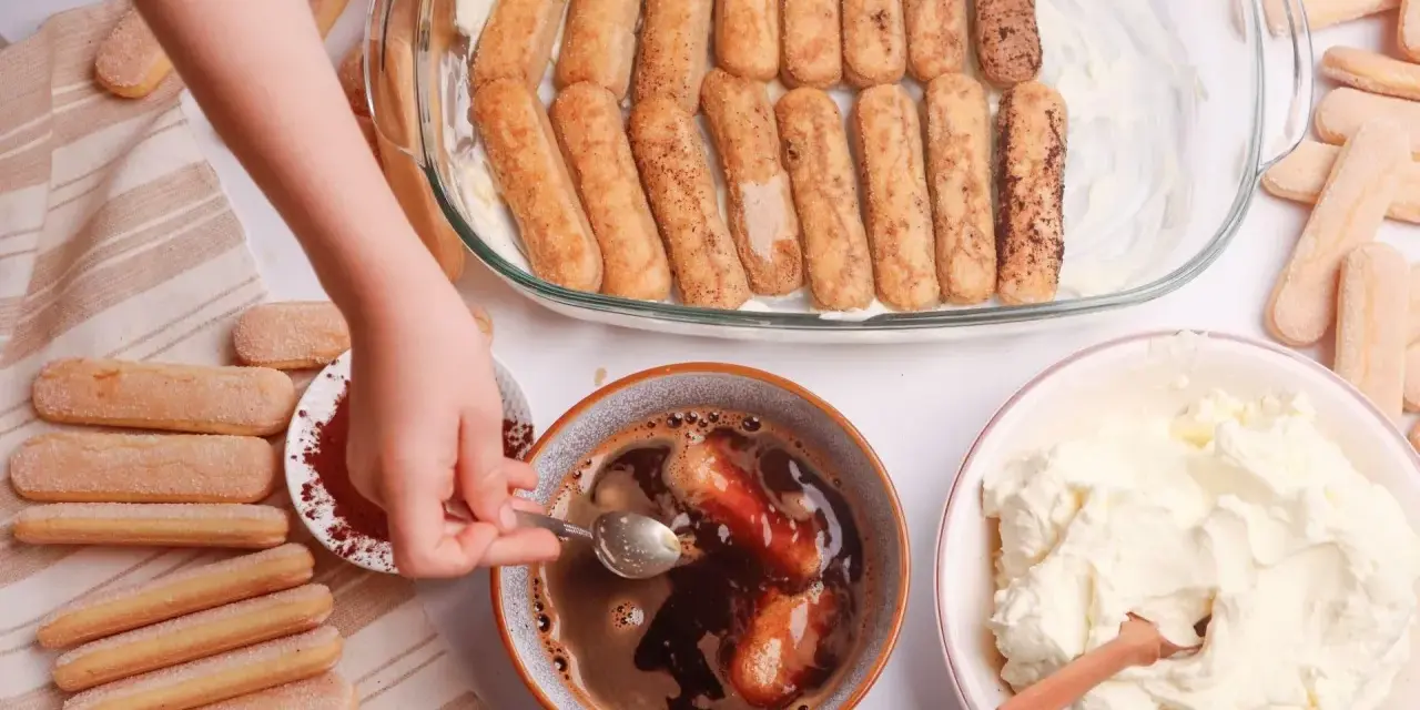 Dziecko zanurza biszkopty w kawie, przygotowując warstwy do tiramisu. Obok krem i kakao.
