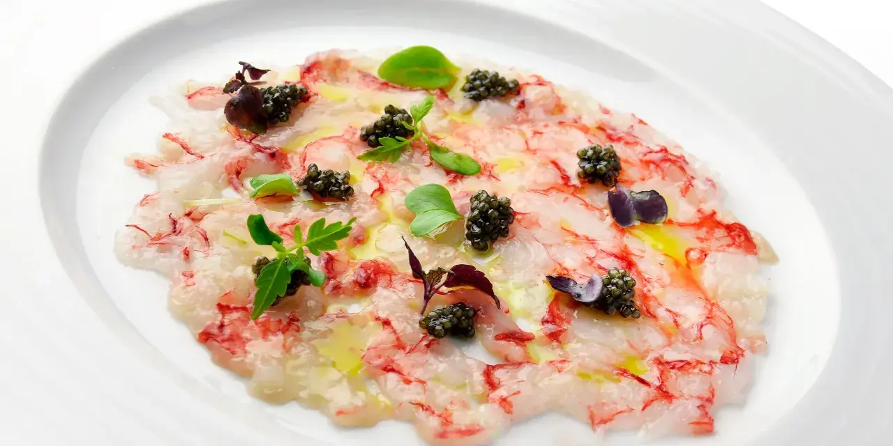 Delicado carpaccio de gambas con caviar negro, aceite de oliva y brotes verdes, presentado en un plato blanco.