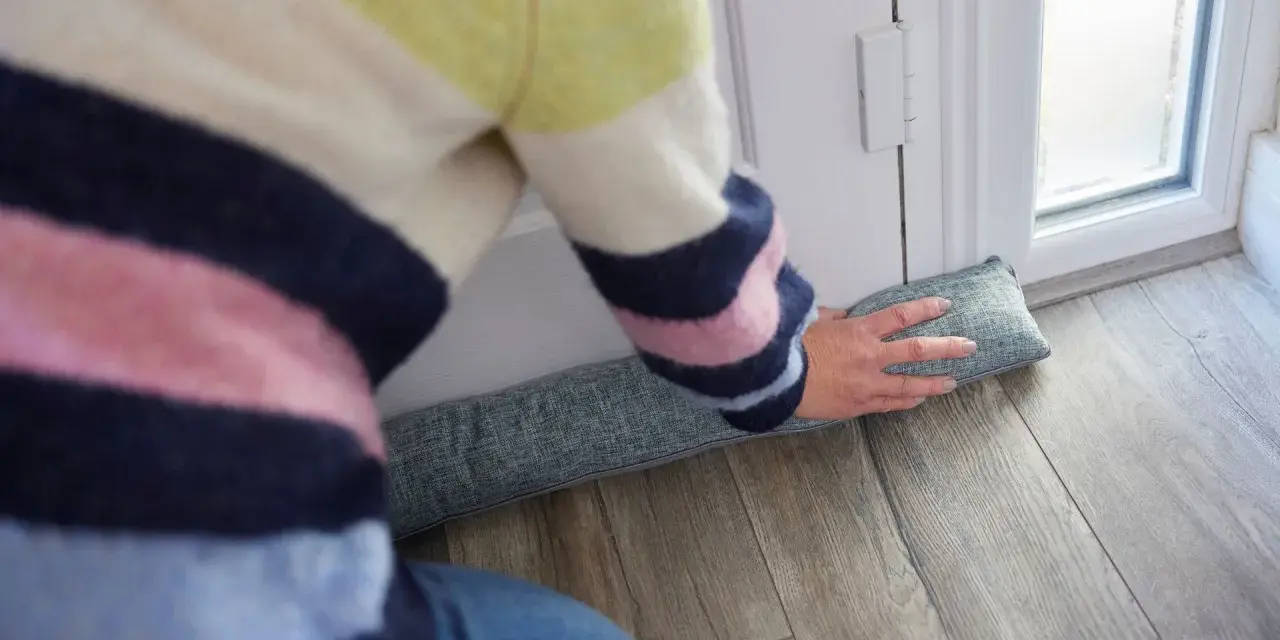 Ocieplanie drzwi wejściowych: osoba w kolorowym swetrze układa podkładkę izolacyjną pod drzwiami, by zatrzymać przeciągi.