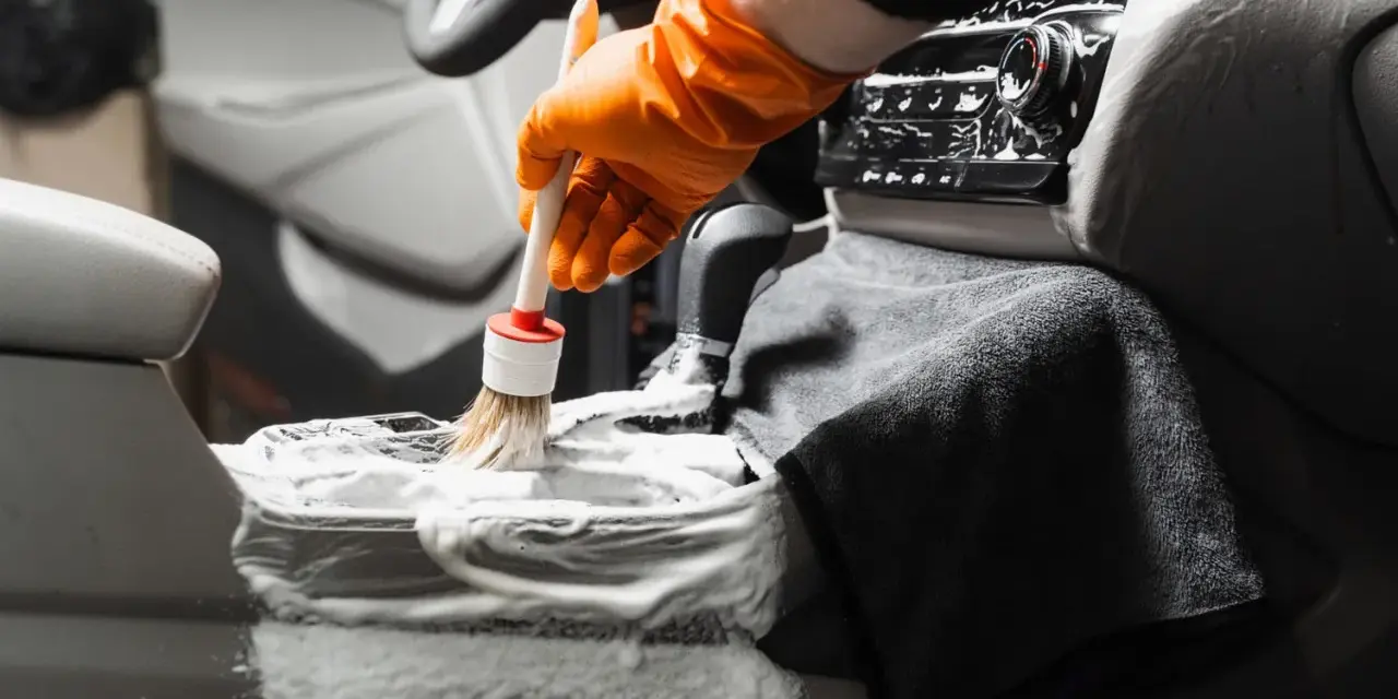 En hand med orange handske använder en **detail brush** för att rengöra bilens mittkonsol med skum.