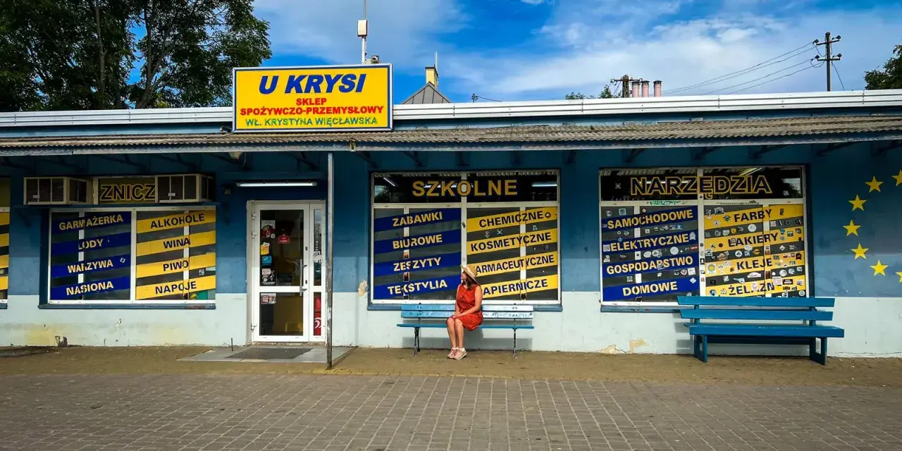 Kobieta w czerwonej sukience siedzi na ławce przed sklepem "U Krysi", na którym widnieją napisy "Szkolne" i "Narzędzia".