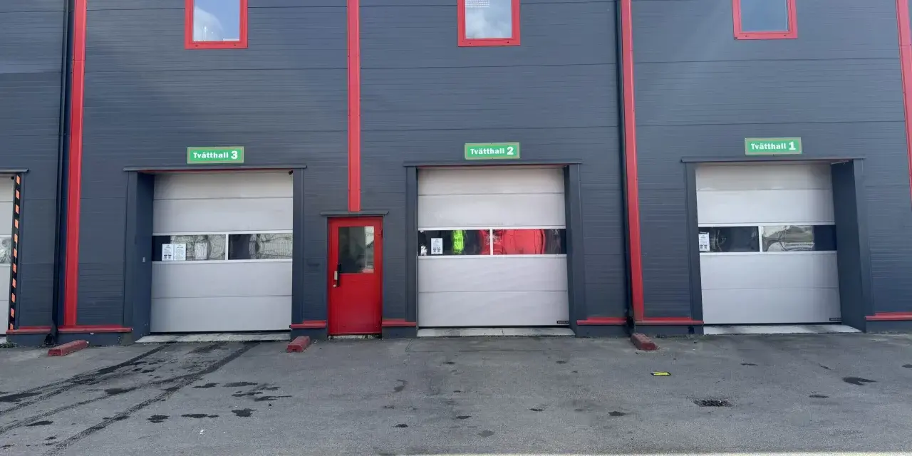 Grå fasad med röda detaljer och tre portar märkta Tvätthall 1, 2 och 3. En röd dörr finns mellan portarna.