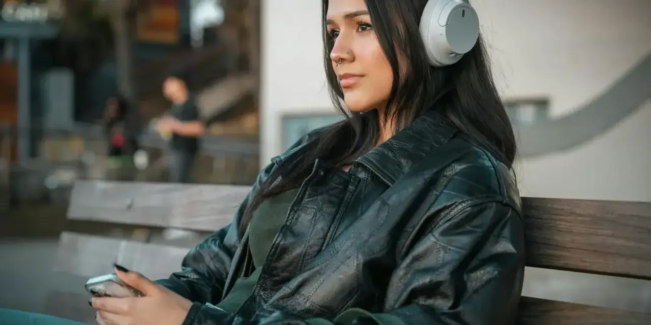 Młoda kobieta w słuchawkach, słuchająca podcastu o wymowie francuskiej, siedzi na ławce, trzymając telefon.