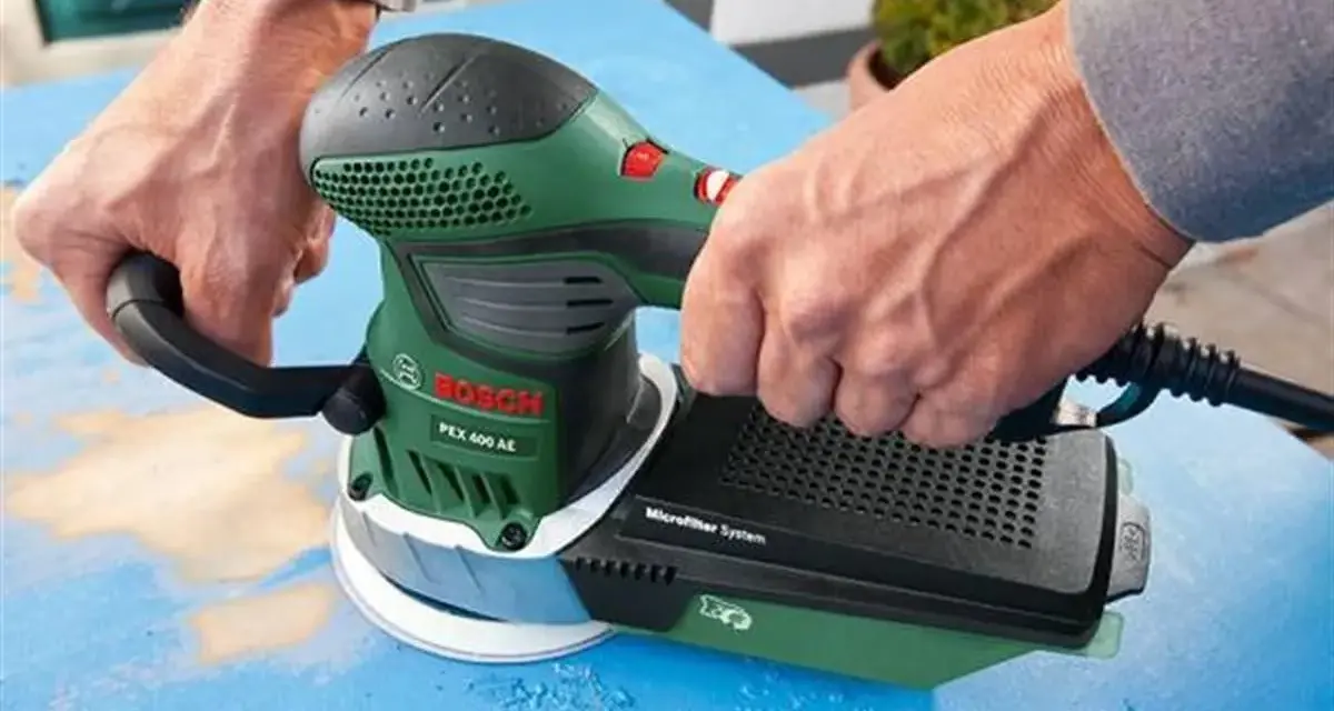 Dłonie trzymają zieloną szlifierkę Bosch PEX 400 AE. To szlifierka oscylacyjna a mimośrodowa, która usuwa starą farbę z niebieskiej powierzchni.