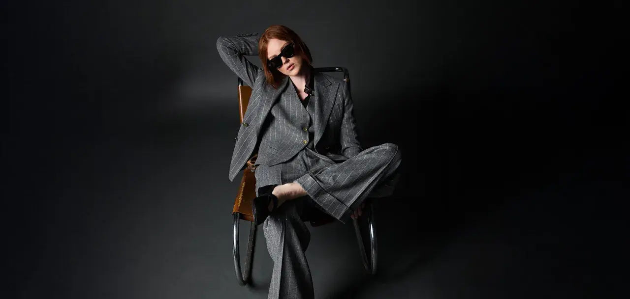 Donna con outfit giacca doppiopetto donna a righe grigie, occhiali da sole, seduta su una poltrona di pelle.