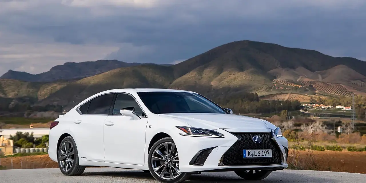 Biały Lexus ES 300h zaparkowany na tle górskiego krajobrazu. Elegancki sedan prezentuje się okazale na tle natury.