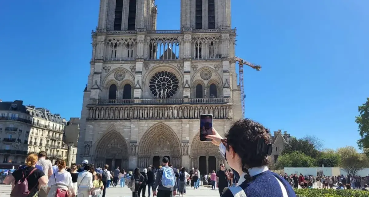 Una persona toma una foto de la fachada de Notre Dame. Mucha gente pasea por la plaza, disfrutando de su visita a Notre Dame.
