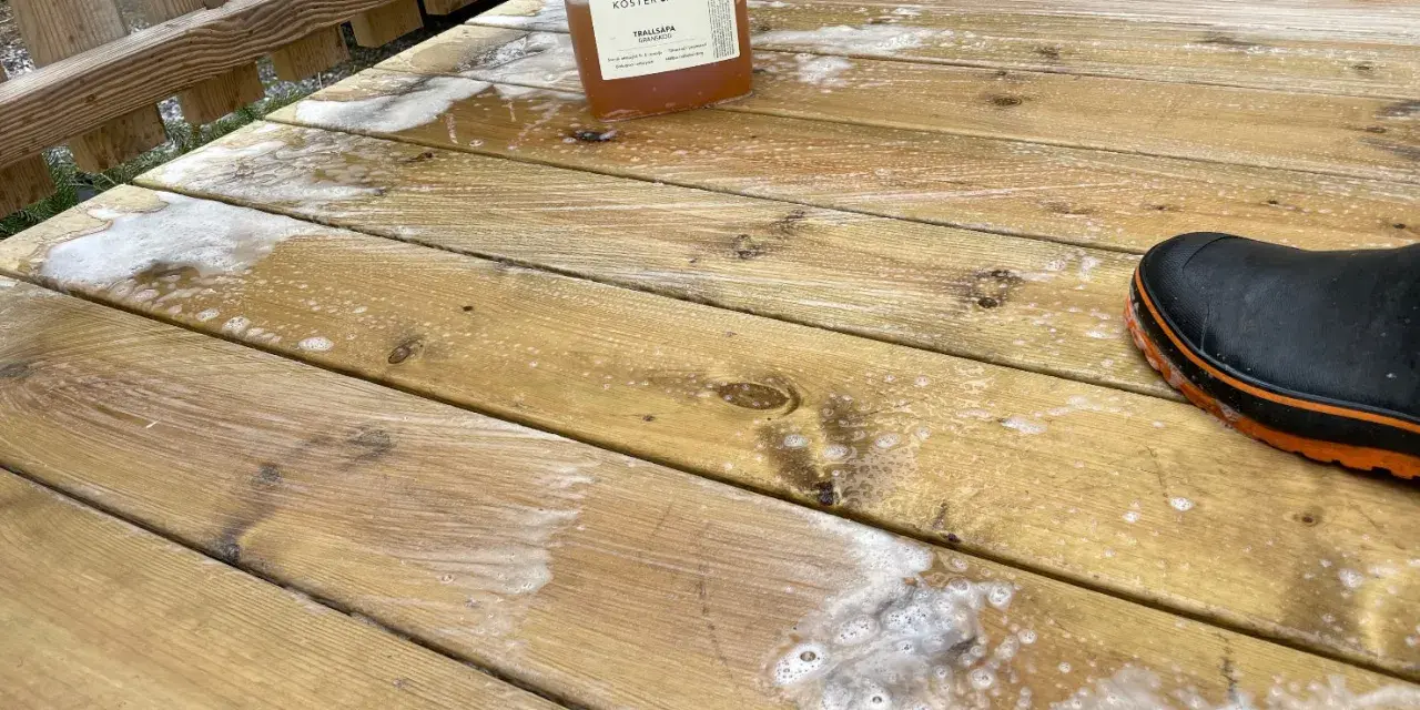 En flaska linoljesåpa står på en altan som håller på att rengöras. En svart stövel med orange kant syns i nedre högra hörnet.