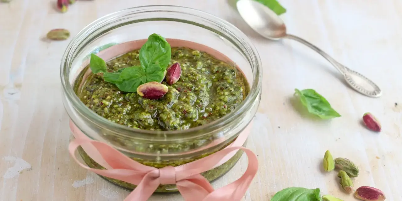 Un vasetto di pesto di pistacchi, decorato con un nastro rosa e guarnito con foglie di basilico e pistacchi interi, pronto per essere gustato.