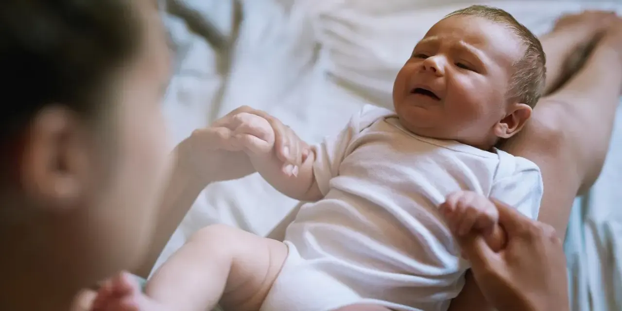 Płaczliwe niemowlę w białym body, trzymające mamę za rękę. Co robić, gdy pojawia się histeria u niemowlaka?
