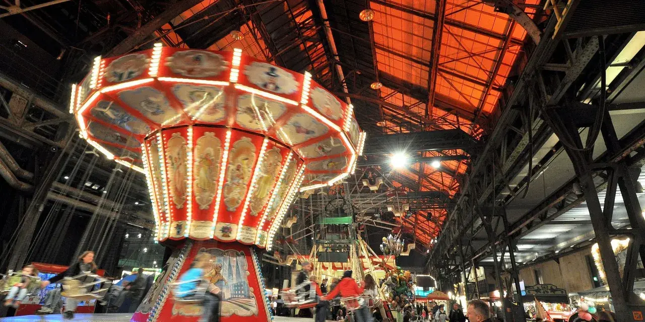 Drehende Kettenkarussells und bunte Lichter auf einem historischen Jahrmarkt in einer alten Industriehalle.