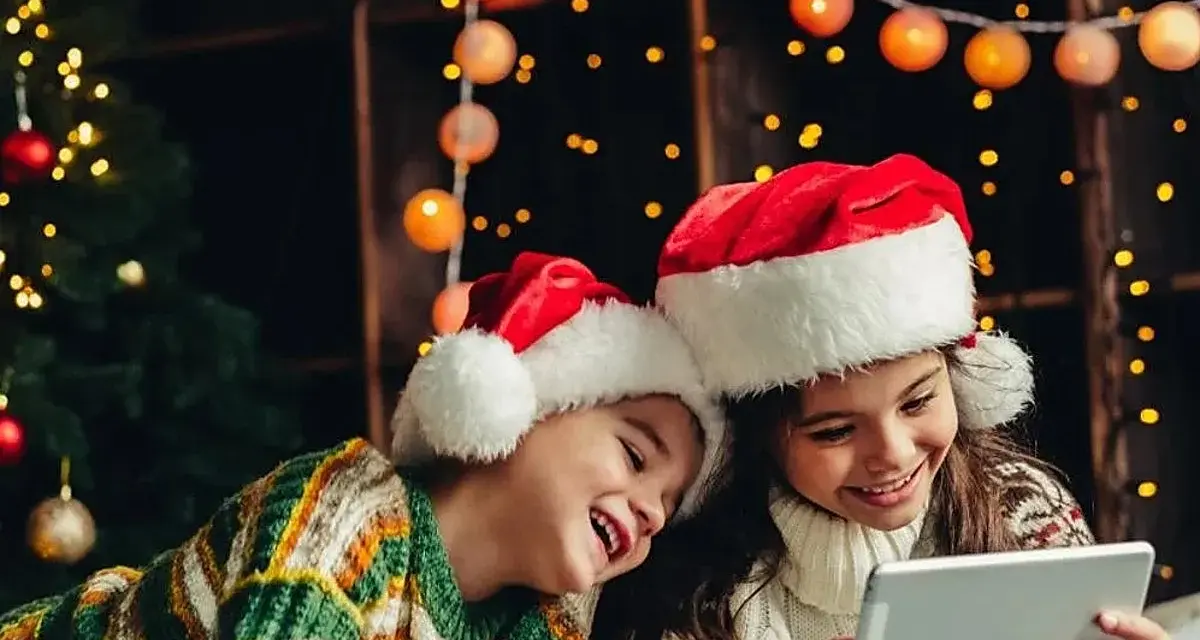 Dos niños con gorros de Santa Claus disfrutan de la tablet, felices cuando dan vacaciones de Navidad, rodeados de luces festivas y un árbol.