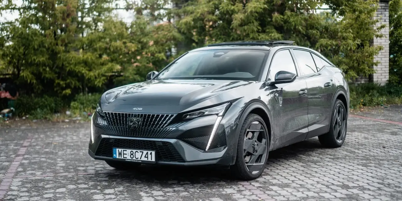 Nowoczesny Peugeot 408 Hybrid w ciemnoszarym kolorze, zaparkowany na brukowanym podjeździe, otoczony zielenią.