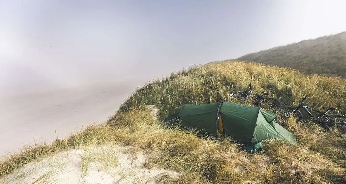 Grön tält och cyklar på en gräsbevuxen sanddyn vid havet. En idyllisk plats för äventyr i sanddynerna Danmark.