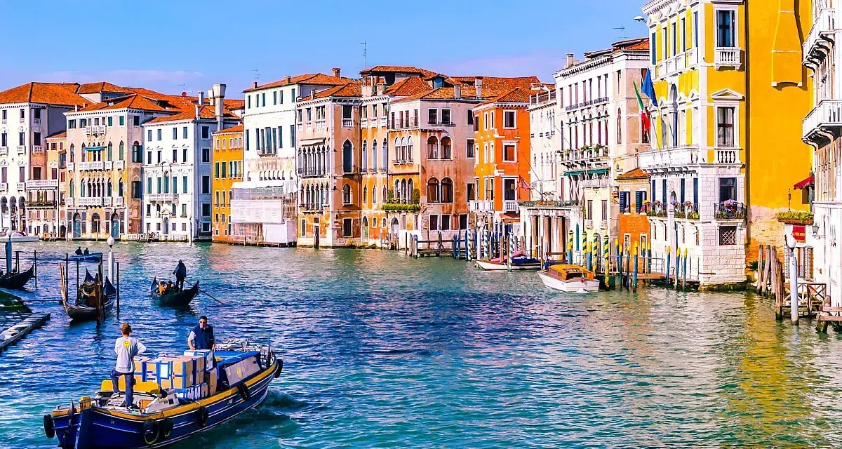 Venezia: turisti in gondola e vaporetti sul Canal Grande, tra palazzi colorati e acque azzurre.