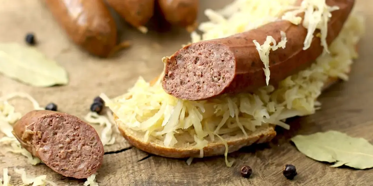 Lungenwurst zubereiten: Eine Scheibe Brot mit Sauerkraut und Lungenwurst belegt, daneben weitere Würste, Gewürze und Lorbeerblätter auf Holzuntergrund.