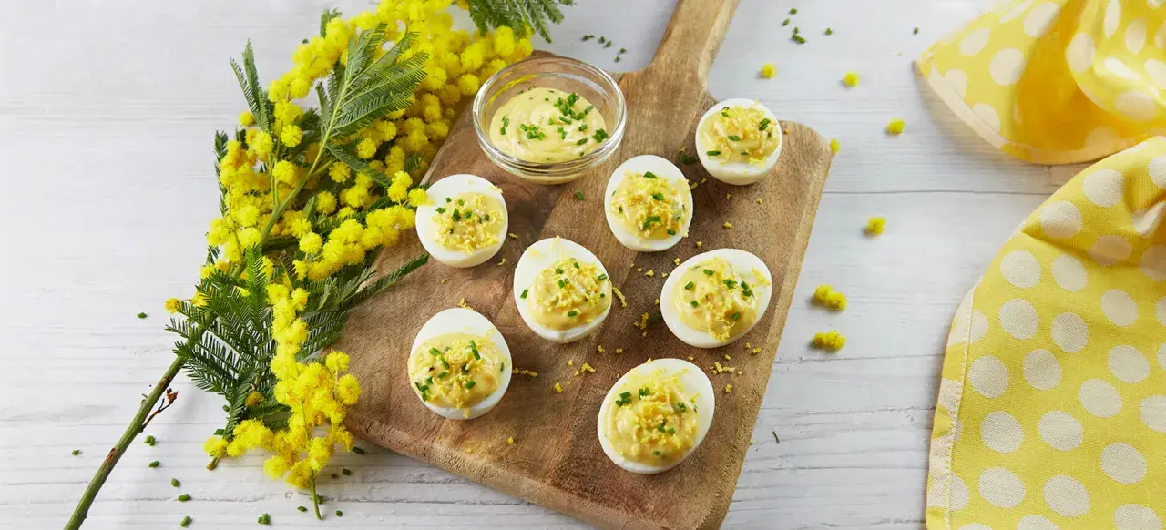 Œufs mimosa garnis d'une sauce crémeuse et de ciboulette, présentés sur une planche en bois avec des fleurs de mimosa. Une recette printanière.