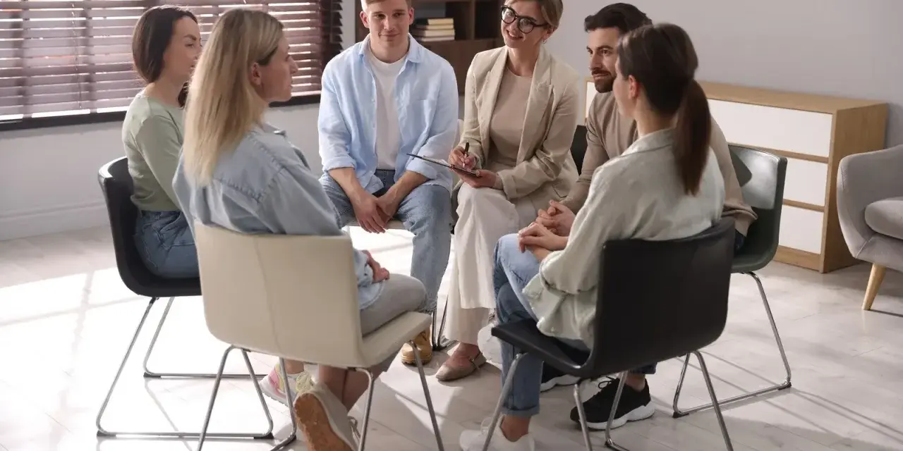 Grupa ludzi siedzi w kręgu, słuchając terapeuty. To scena z ośrodka dla uzależnionych, gdzie wsparcie i zrozumienie pomagają w powrocie do zdrowia.