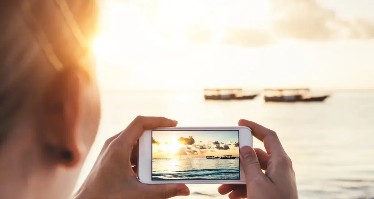 Une personne prend une photo d'un coucher de soleil sur la mer avec des bateaux au loin. Une belle photo à faire à la plage.