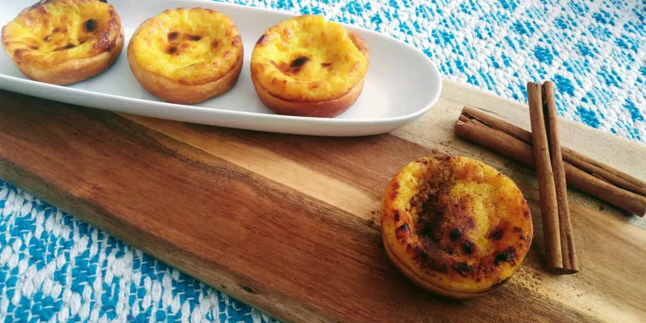 Vier goldbraune, karamellisierte Pastéis de Nata auf einem weißen Teller, daneben Zimtstangen auf einem Holzbrett.