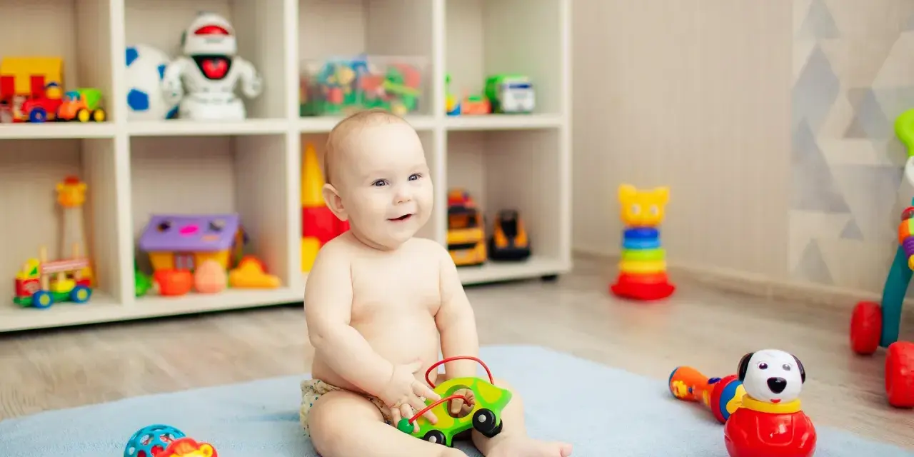 Uśmiechnięte, 8-miesięczne dziecko nie siedzi, bawi się zielonym autkiem na niebieskim kocyku. W tle regał z zabawkami.