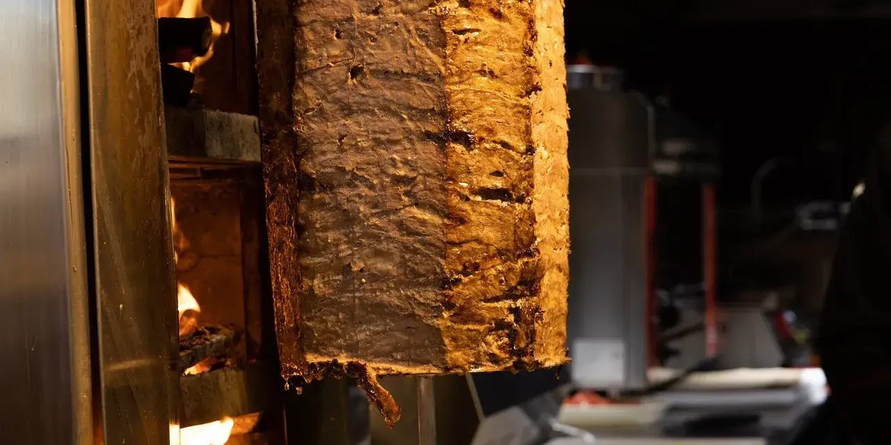 Duży pionowy obrotowy grill z mięsem, obok płomienie ognia.