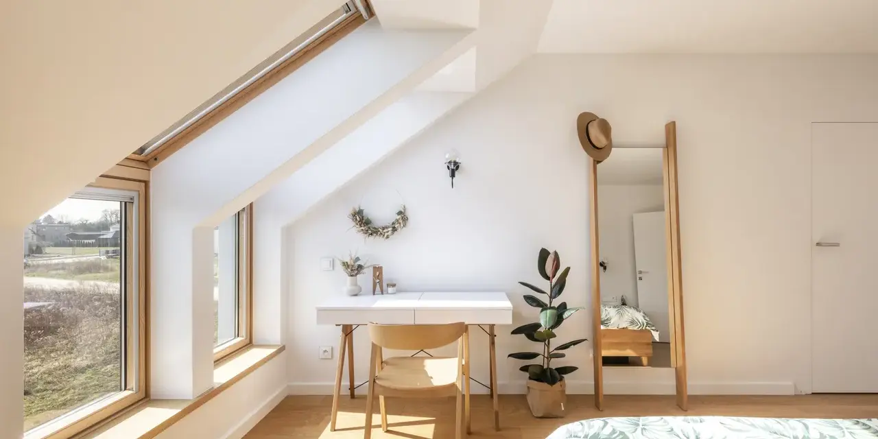 Angolo studio mansardato inondato da la luce più naturale. Scrivania, sedia in legno, specchio alto e pianta.