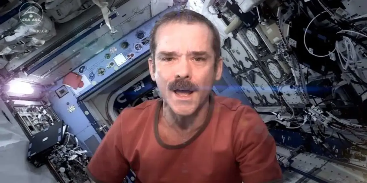 Chris Hadfield singt "Space Oddity" in der ISS. Sein Gesicht mit Schnurrbart ist im Vordergrund, der Raumschiff-Innenraum im Hintergrund.
