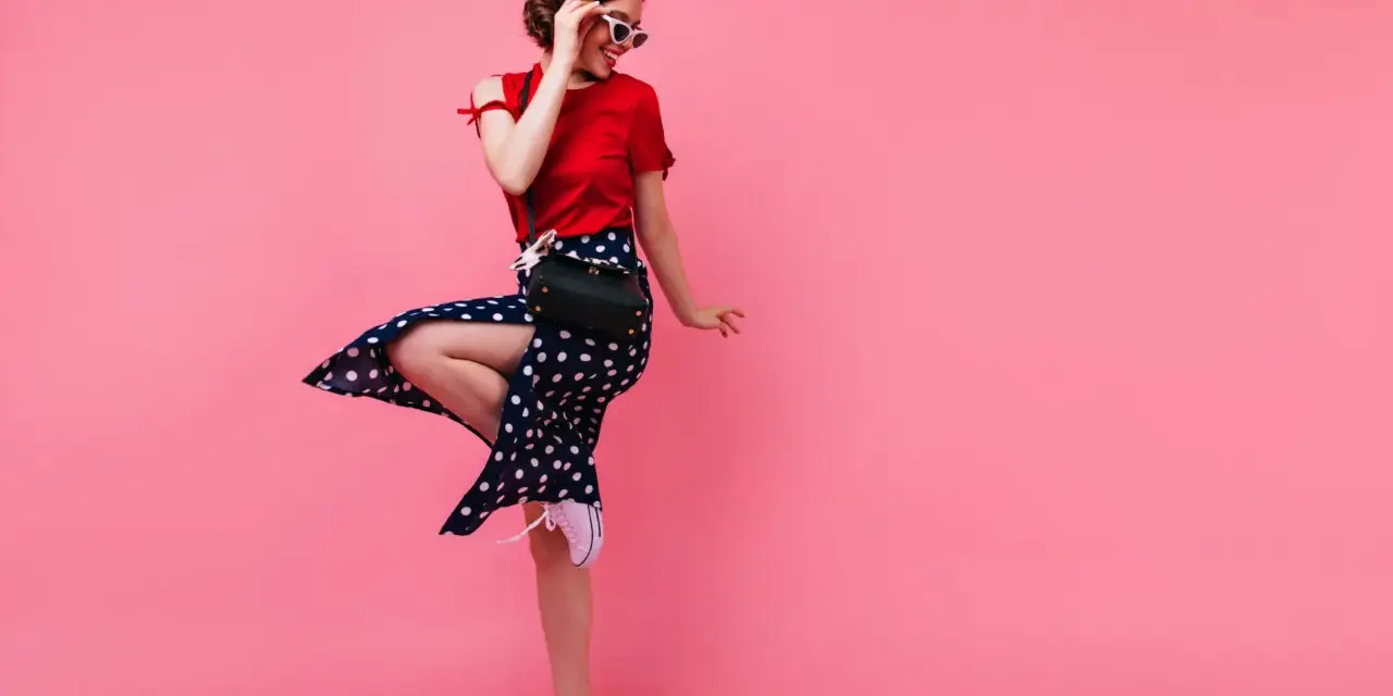 Kobieta w stylu pin-up w czerwonym berecie i sukience w grochy skacze na różowym tle.