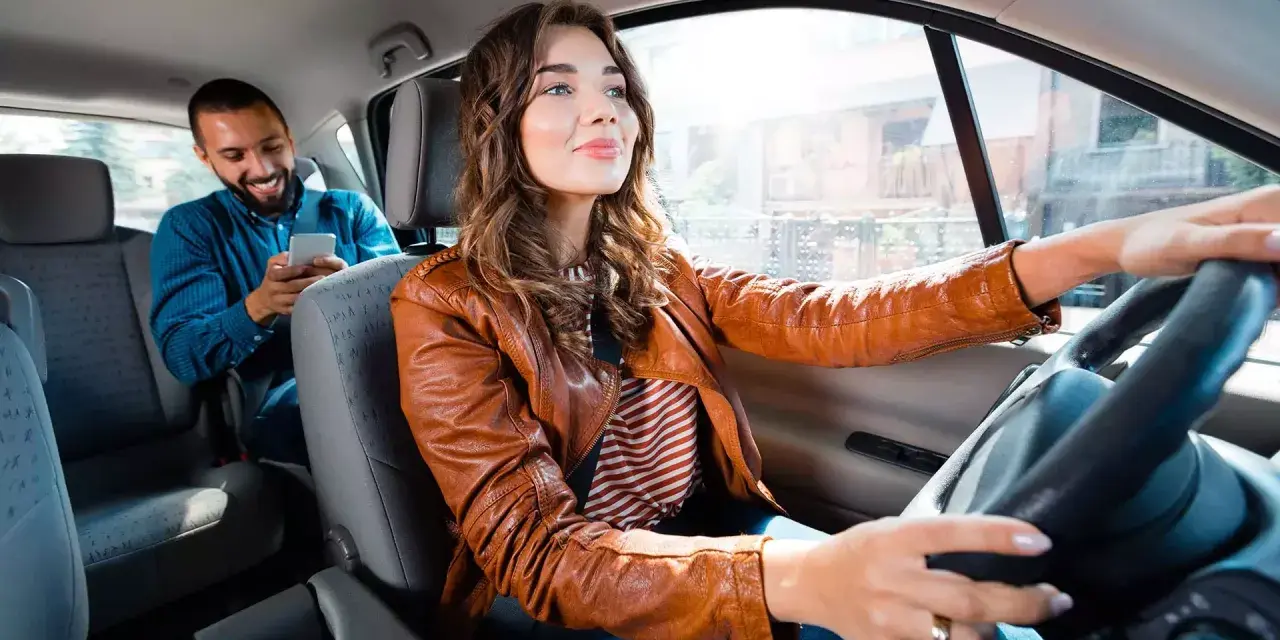 Fahrerin im Taxi, ihr Fahrgast schaut aufs Handy. Sie hat ihren Taxi Schein gut sichtbar.