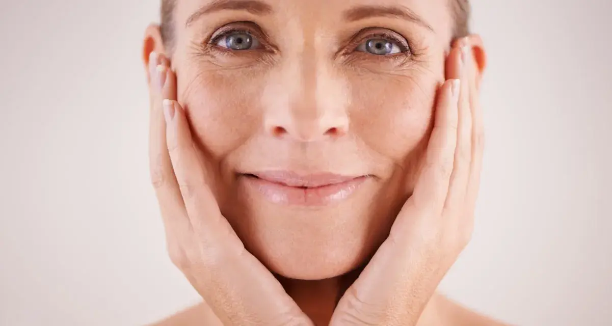 Visage d'une femme aux mains sur les joues, montrant les signes du temps et les défis des problèmes de peau et ménopause.